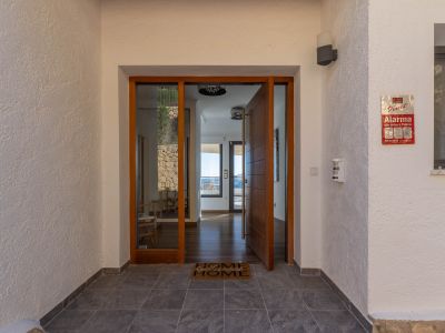 Hoofdingang met houten draaideur en glazen paneel, die een licht interieur met houten vloeren en uitzicht op zee onthult. Gestructureerde buitenmuren en donkere tegelvloer.