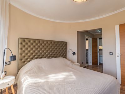 Hoofdslaapkamer met gestoffeerd hoofdbord, een kenmerkende gebogen muur en natuurlijk licht gefilterd door transparante gordijnen, aangevuld met houten deuren.
