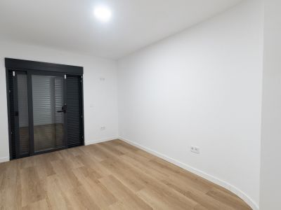 Lichte kamer met lichte houtnerf laminaatvloer en witte muren. Een donkergrijze glazen schuifdeur met geïntegreerde jaloezieën biedt toegang tot buiten.