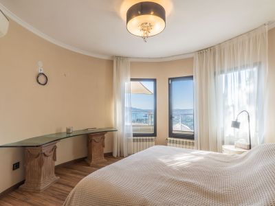 Lichte slaapkamer met panoramisch zeezicht door grote ramen met voile gordijnen en een uniek glazen bureau op gesneden houten sokkels.