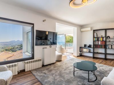 Lichte woonkamer met panoramisch uitzicht op de bergen vanuit het raam en toegang tot het terras met zeezicht. Voorzien van een houten vloer, een groot grijs vloerkleed en een moderne boekenkast.