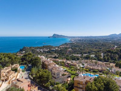 Luchtfoto van de kustlijn van Altea Hills, met de Middellandse Zee, de Peñón de Ifach en de skyline van Benidorm in de verte. Terracotta daken en zwembaden zijn zichtbaar tussen weelderig groen.