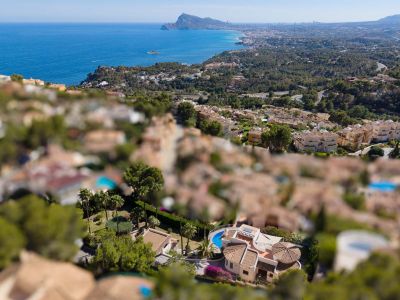 Luchtfoto van een villa in Altea Hills met een gebogen zwembad, terracotta daken en weelderige tuinen, met de Middellandse Zee en kustlijn op de achtergrond.