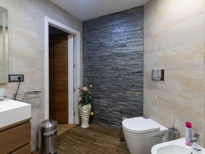 Moderne badkamer met donkere gestapelde stenen accentmuur, lichtbeige tegels en houtlookvloer. Voorzien van toilet, bidet en badmeubel met wastafel.