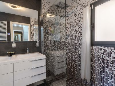 Moderne badkamer met dubbele wastafel, wit meubel en verlichte spiegel. De glazen douchewand heeft mozaïektegels in grijstinten en een geïntegreerd raam.