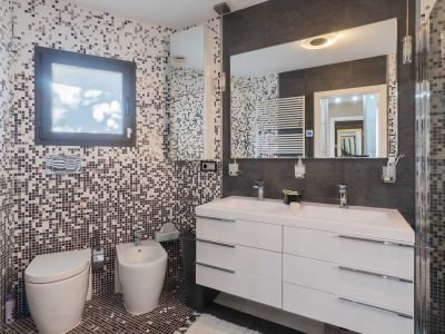 Moderne badkamer met mozaïektegels op wanden en vloer in aardse/grijze tinten. Beschikt over een dubbele zwevende wastafel, grote verlichte spiegel, wandtoilet, bidet en handdoekradiator.
