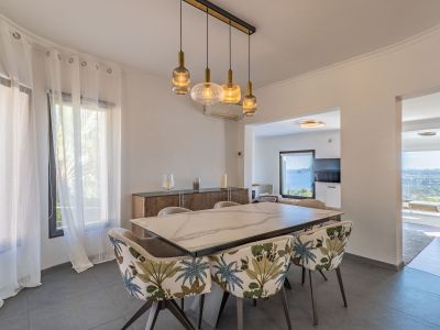 Moderne eetkamer met marmerlook tafel en stoelen met tropisch bladpatroon. Een lineaire kroonluchter verlicht de ruimte, die vanuit aangrenzende kamers panoramisch uitzicht op zee biedt.
