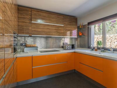 Moderne keuken met glanzende bovenkasten in houtnerf en feloranje onderkasten. Wit aanrechtblad, metallic mozaïek achterwand en een raam met uitzicht op een stenen muur.