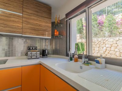 Moderne keuken met wit kwartswerkblad, oranje onderkasten en bovenkasten met houtnerf. Het raam biedt uitzicht op een natuurstenen muur en weelderig groen.