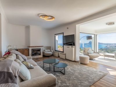 Moderne woonkamer met lichte bank, open haard en grote ramen die een panoramisch uitzicht op zee en stad bieden. Hedendaags meubilair versterkt de lichte ruimte.