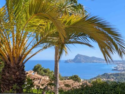 Panoramisch uitzicht op de Middellandse Zee, omlijst door palmbomen, met de daken van Altea Hills en de verre Peñón de Ifach onder een helderblauwe lucht.