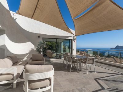 Ruim buitenterras met lounge- en eethoek onder een schaduwdoek, met panoramisch zee- en kustzicht via een glazen balustrade.
