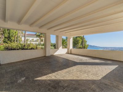 Ruime overdekte carport met gestempelde betonvloer en wit balkenplafond. Het biedt panoramisch uitzicht op de Middellandse Zee en de kustlijn, omgeven door weelderig groen.