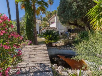 Rustieke houten loopbrug over een serene vijver, omzoomd door levendige roze oleanders en hoge palmbomen, leidend naar de villa onder de mediterrane zon.