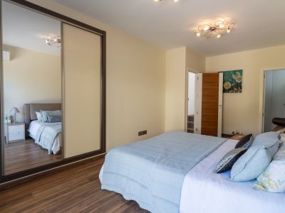 Slaapkamer met bed in lichtblauwe stoffen, houten laminaatvloer en schuifkast met spiegelpaneel. Een moderne plafondlamp en bloemenkunst aan de muur zijn aanwezig.