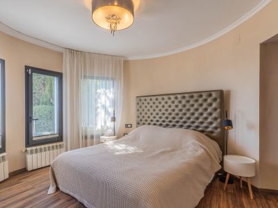 Slaapkamer met groot bed en gecapitonneerd hoofdbord. Gebogen wand met donkeromlijste ramen en lichte gordijnen. Houten vloer, radiatoren en een moderne plafondlamp.