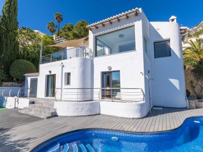 Witte villa-exterieur met grote ramen en terrassen met glazen balustrades. Het blauwe mozaïekzwembad en het grijze composietdek zijn omringd door cipressen en palmbomen.