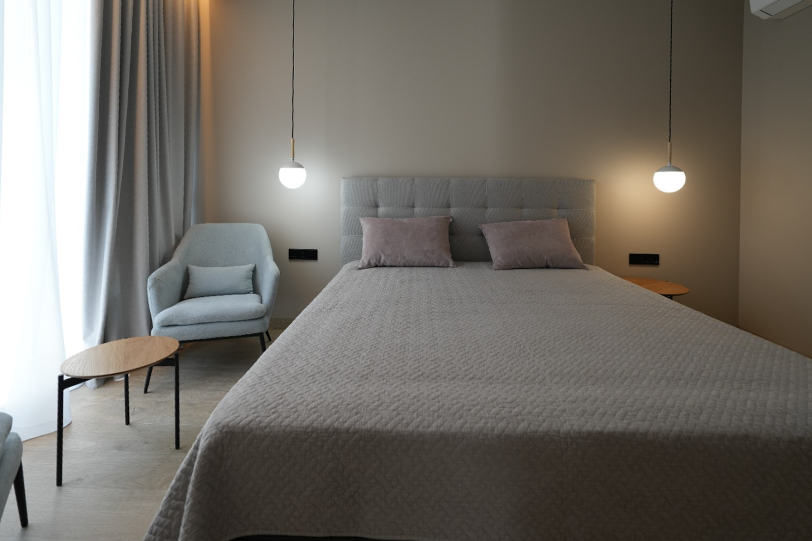 Elegante slaapkamer met een grijs gestoffeerd bed, twee moderne hanglampen en een comfortabele fauteuil. Lichte gordijnen en houtlook vloer creëren een serene sfeer.