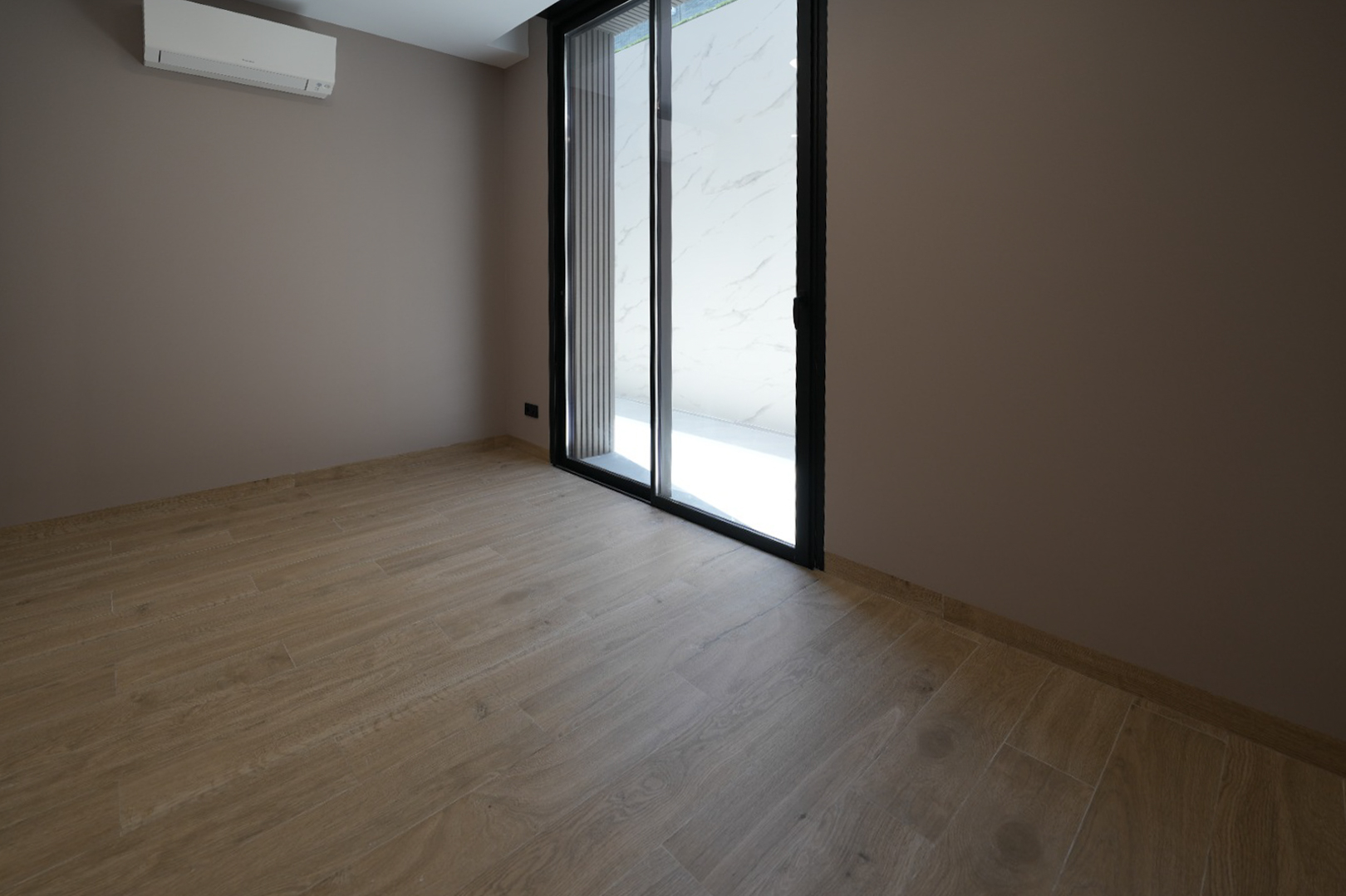 Lege kamer met licht houtlook tegelvloer, taupekleurige muren en een zwarte schuifpui. Een wandgemonteerde airconditioningunit is aanwezig.