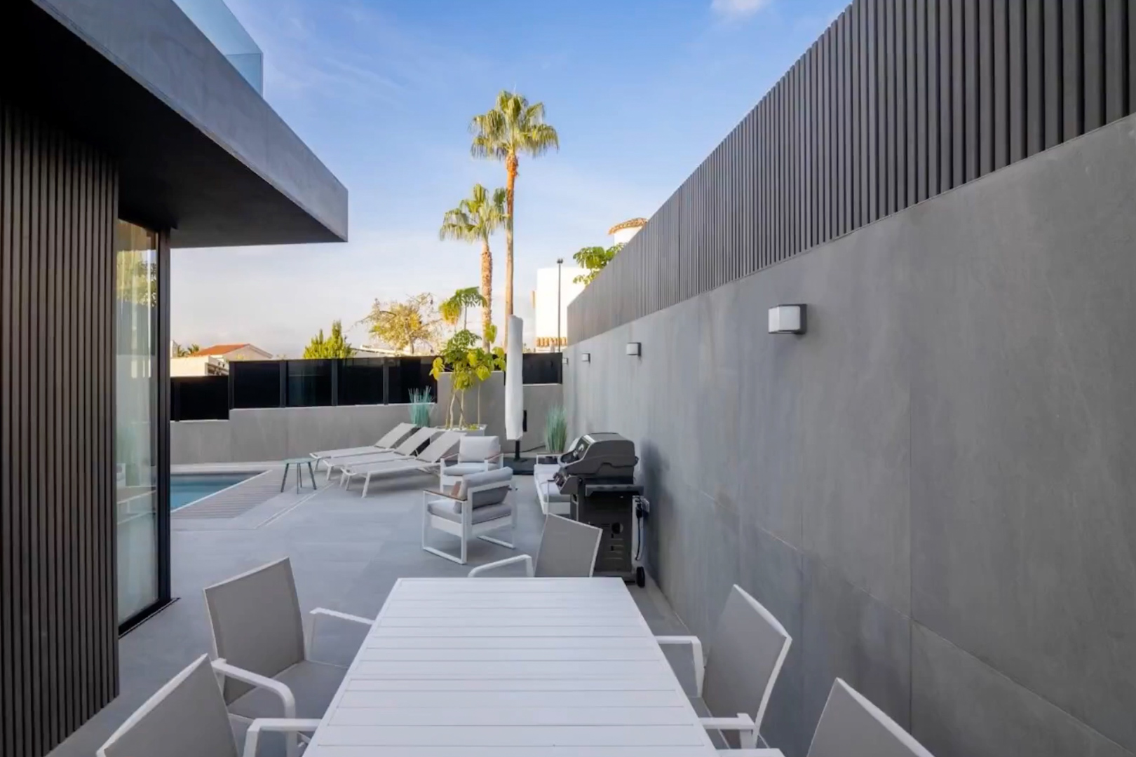Modern buitenterras met witte lamellen eettafel en stoelen, loungehoek en barbecue, omgeven door grijze tegelwanden en donkere privacyschermen, met uitzicht op het zwembad.