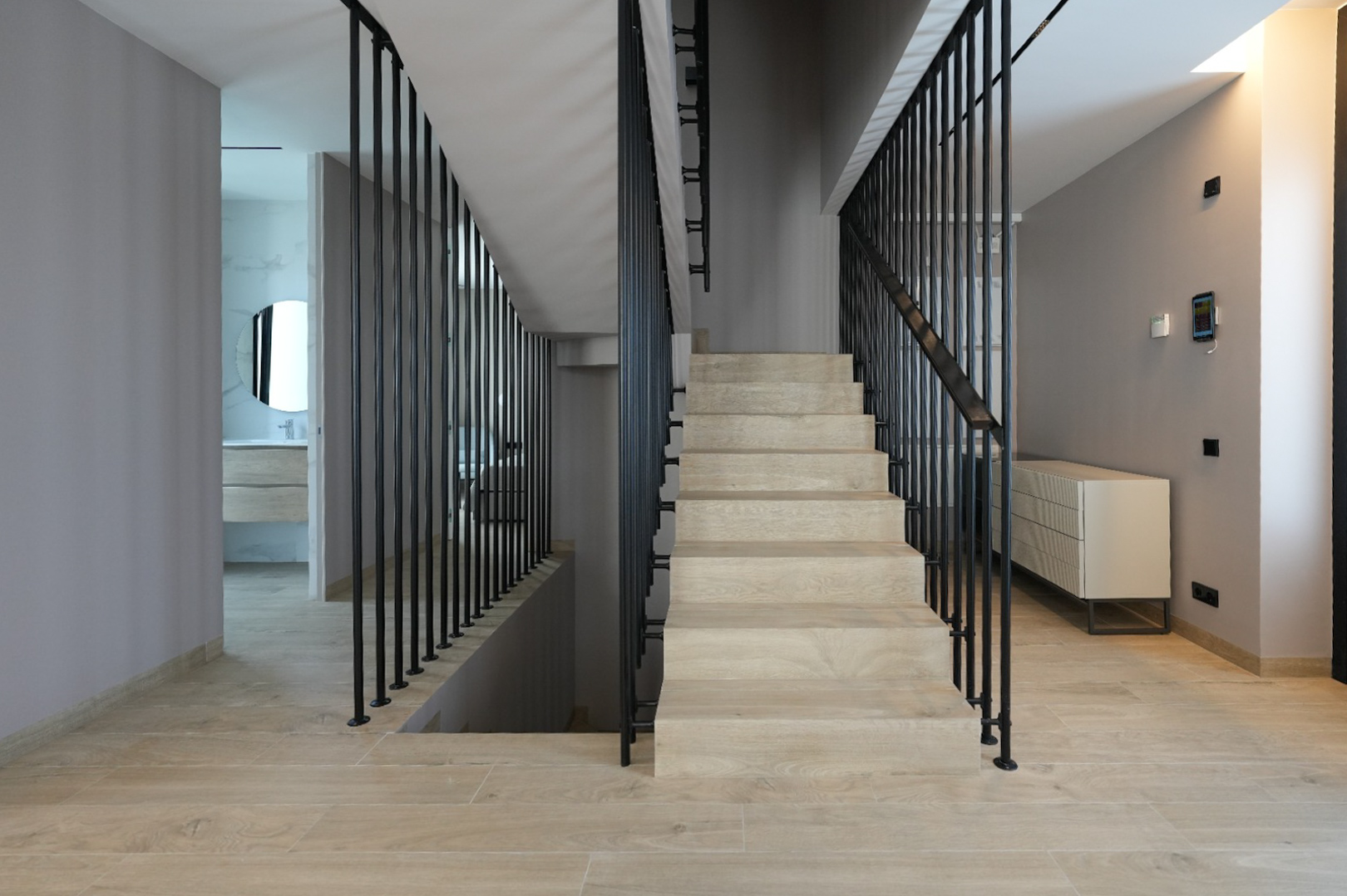 Modern interieur met lichte houtlook trap en strakke zwarte verticale balustrade. Een smart home bedieningspaneel is zichtbaar, leidend naar een badkamer met ronde spiegel.