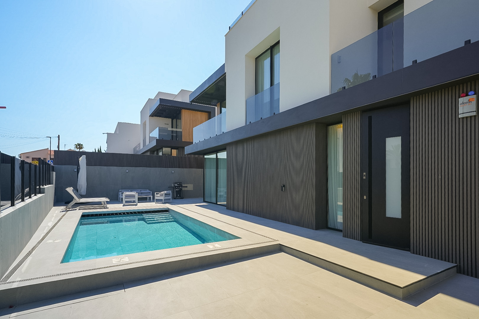 Modern villa-exterieur met rechthoekig zwembad en geïntegreerde treden, omringd door lichte bestrating. De begane grond toont een donkere lamellen gevel en een opvallende toegangsdeur.