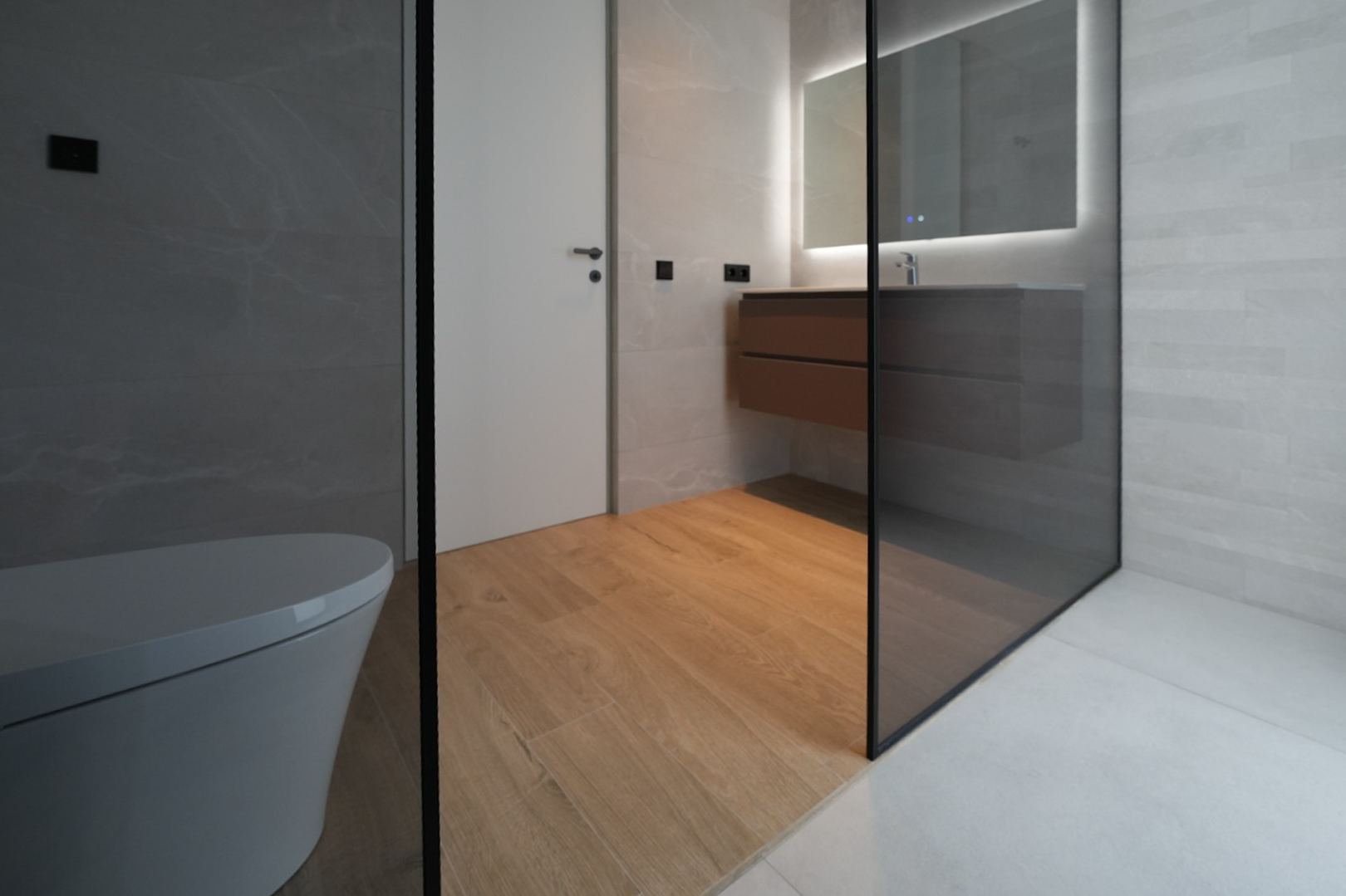 Moderne badkamer met zwevend toilet, houten wastafelmeubel, verlichte spiegel en glazen douchewand. Houtlook vloertegels contrasteren met de grijze wand- en douchevloertegels.