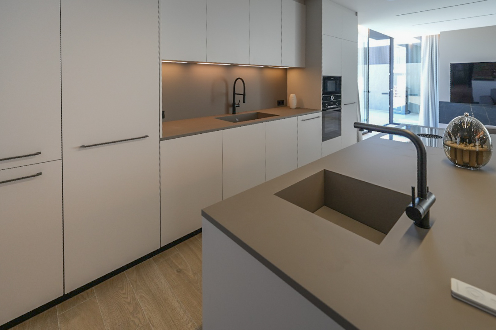 Moderne keuken met kookeiland en dubbele geïntegreerde spoelbak. Minimalistische kasten in neutrale tinten, inbouwapparatuur en lichte houten vloer, met toegang naar buiten.