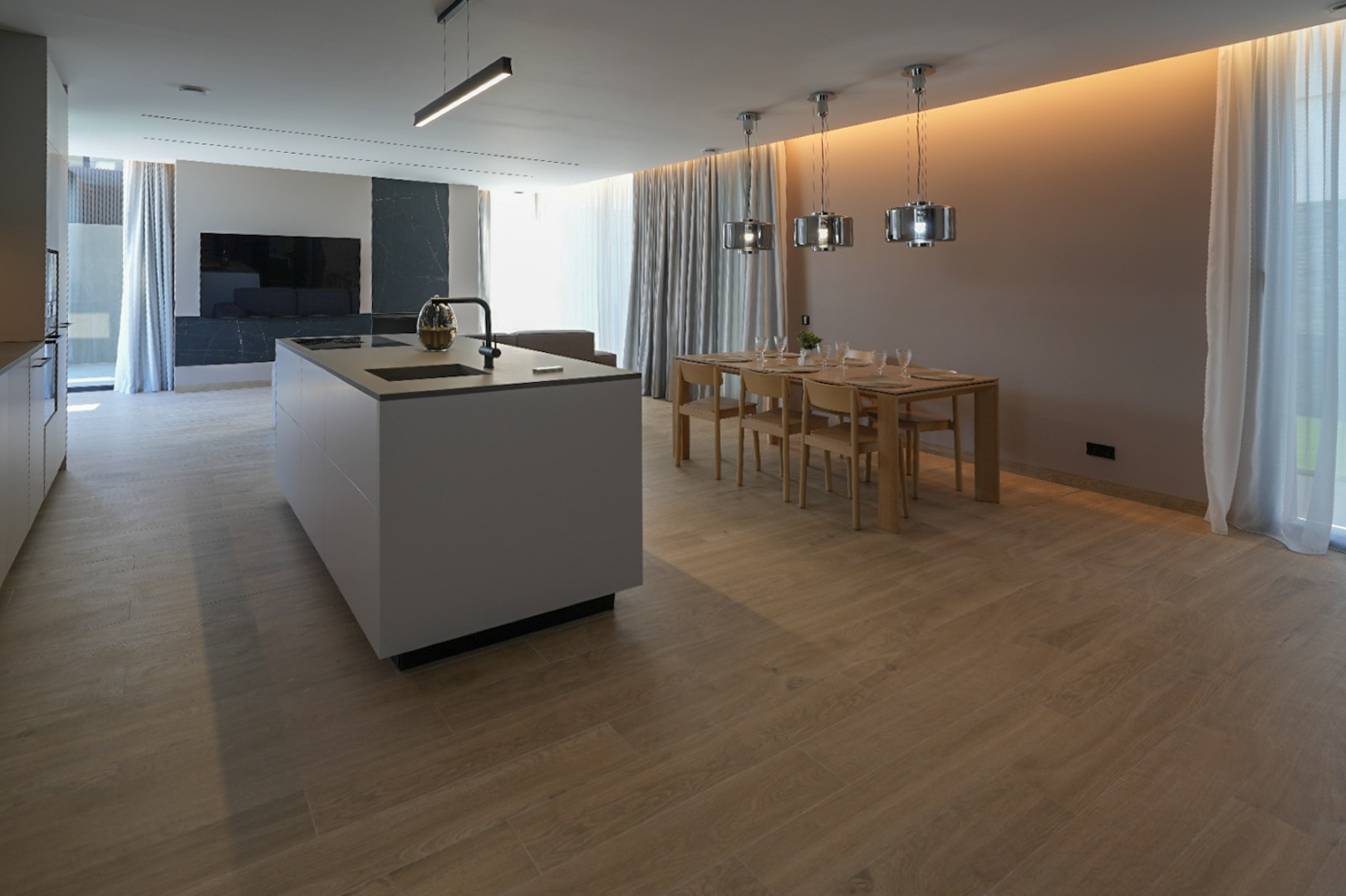 Moderne keukeneiland met geïntegreerde spoelbak en kookplaat, grenzend aan een eetruimte met houten tafel en eigentijdse hanglampen. Lichtgrijze houtlook tegelvloer.