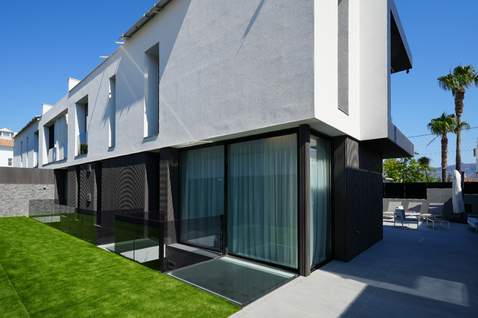 Moderne villa-exterieur met contrasterende wit-grijze en donkere gevels, grote glazen schuifdeuren, kunstgrasgazon en een betegeld terras met buitenmeubilair.