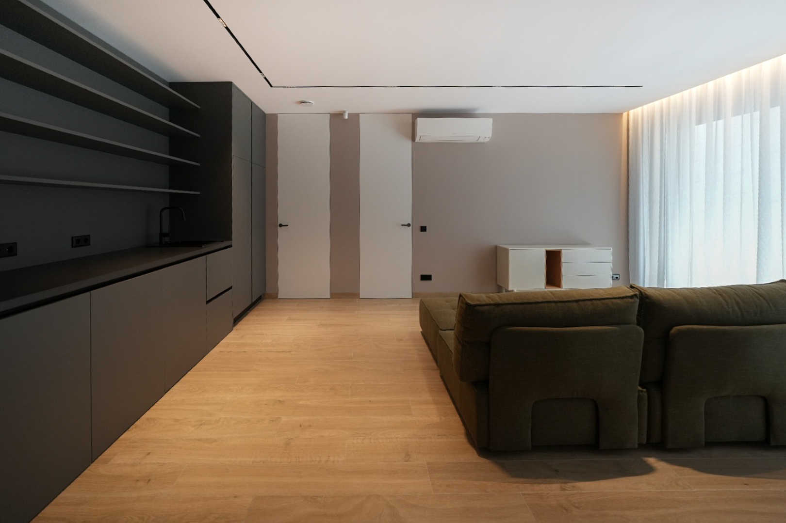 Moderne woonruimte met donkergrijze geïntegreerde kitchenette, lichte houtlook vloer en groene modulaire bank. Vlakke deuren en natuurlijk licht.