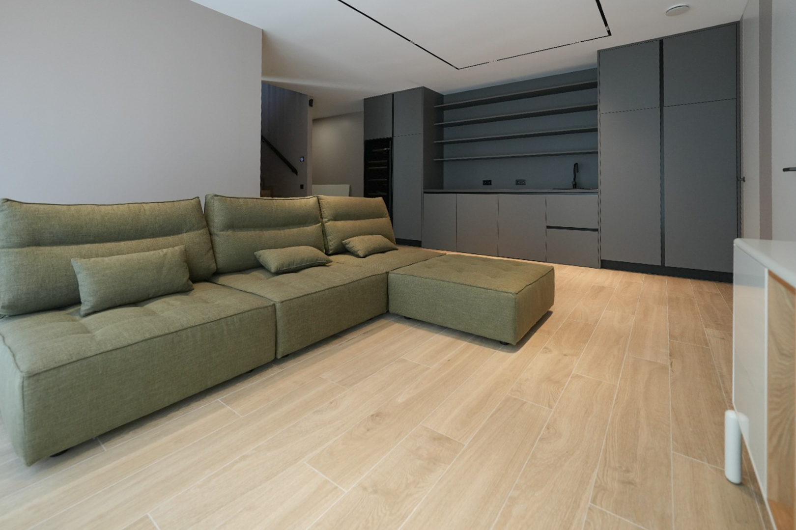 Olijfgroene modulaire bank in open woonruimte met licht houtlook tegelvloer. Donkergrijze inbouwkasten met spoelbak en planken op de achtergrond.