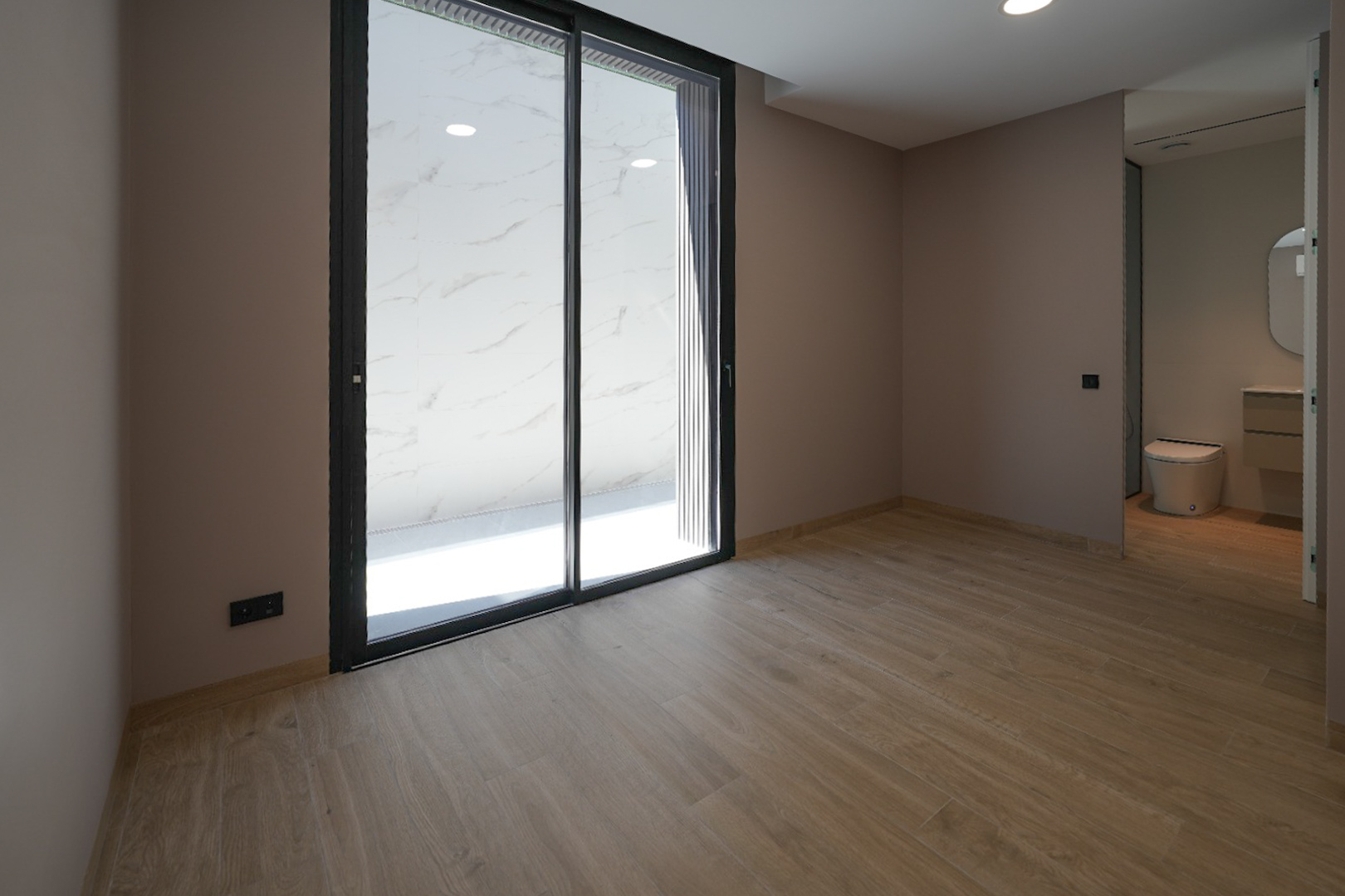 Ruime kamer met houtlook tegelvloer en lichte muren. Grote zwart omkaderde glazen schuifdeuren openen naar een buitenruimte met witte marmerlook tegels. Geïntegreerde badkamer zichtbaar.