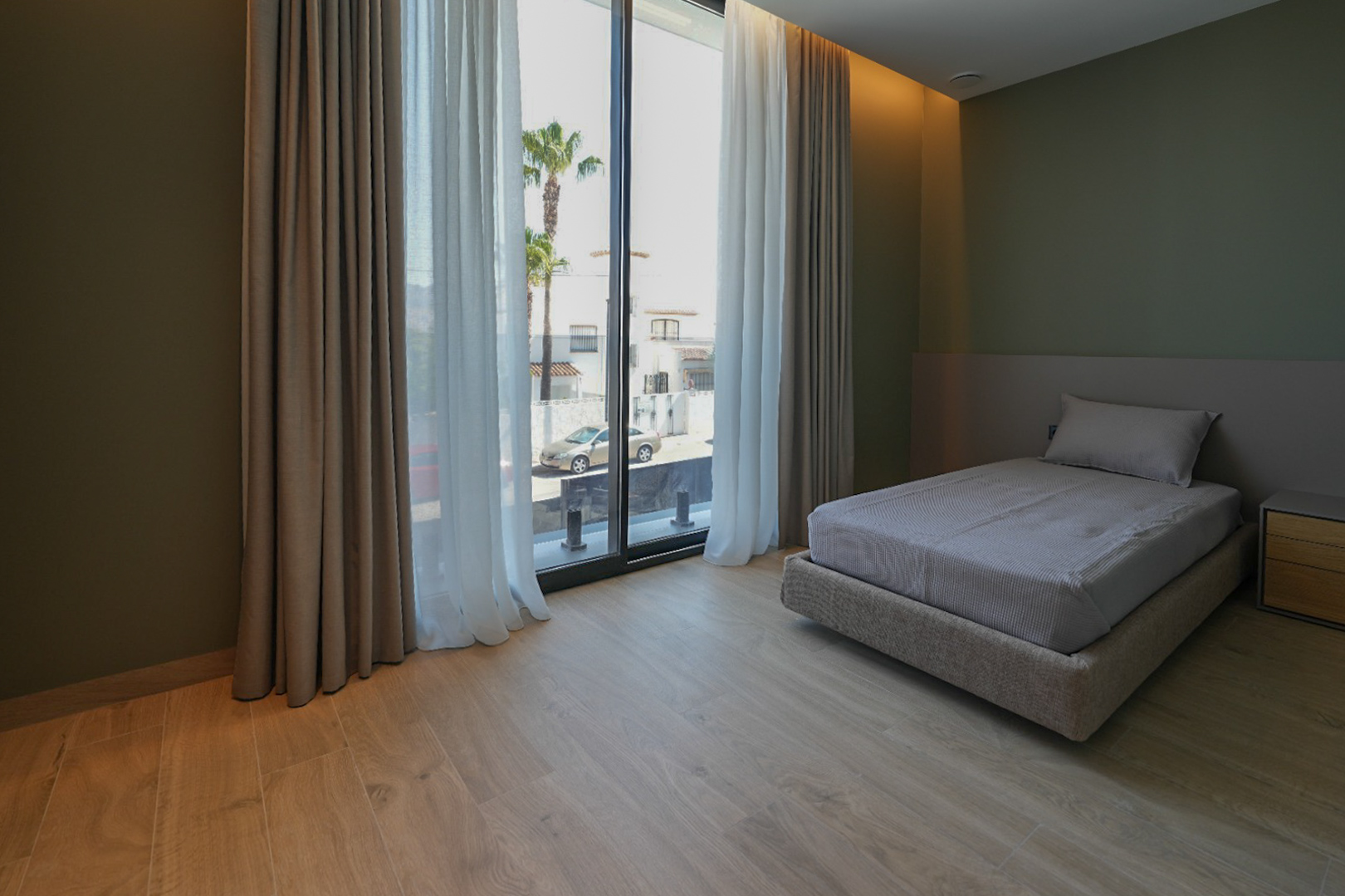 Slaapkamer met eenpersoonsbed, lichte houtlook vloer en olijfgroene muren. Grote ramen met dubbele gordijnen bieden uitzicht op de straat en palmbomen.