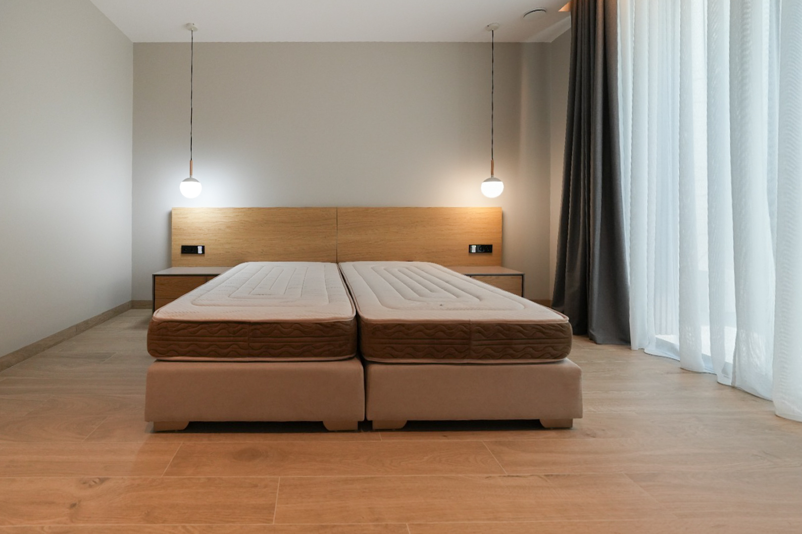 Slaapkamer met tweepersoonsbed op gestoffeerde bedframes, licht houten hoofdbord met geïntegreerde nachtkastjes en twee moderne hanglampen. Groot raam met gordijnen.