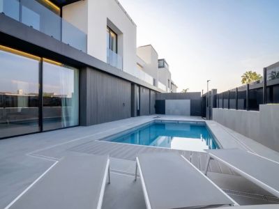 Blauw buitenzwembad, omringd door een grijze tegelpatio en composiet vlonder. De moderne gevel met grote glazen schuifdeuren weerspiegelt de heldere lucht.