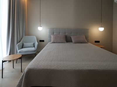 Elegante slaapkamer met een grijs gestoffeerd bed, twee moderne hanglampen en een comfortabele fauteuil. Lichte gordijnen en houtlook vloer creëren een serene sfeer.