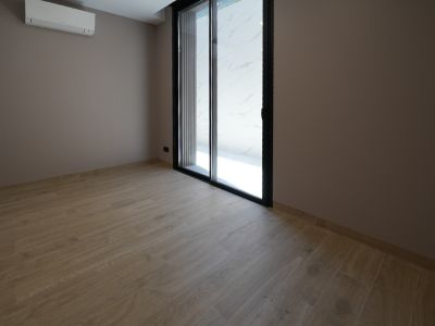 Lege kamer met licht houtlook tegelvloer, taupekleurige muren en een zwarte schuifpui. Een wandgemonteerde airconditioningunit is aanwezig.