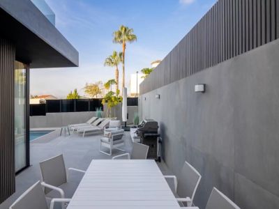 Modern buitenterras met witte lamellen eettafel en stoelen, loungehoek en barbecue, omgeven door grijze tegelwanden en donkere privacyschermen, met uitzicht op het zwembad.