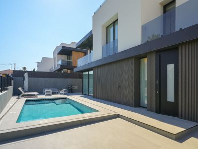 Modern villa-exterieur met rechthoekig zwembad en geïntegreerde treden, omringd door lichte bestrating. De begane grond toont een donkere lamellen gevel en een opvallende toegangsdeur.