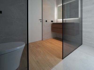 Moderne badkamer met zwevend toilet, houten wastafelmeubel, verlichte spiegel en glazen douchewand. Houtlook vloertegels contrasteren met de grijze wand- en douchevloertegels.