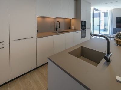 Moderne keuken met kookeiland en dubbele geïntegreerde spoelbak. Minimalistische kasten in neutrale tinten, inbouwapparatuur en lichte houten vloer, met toegang naar buiten.