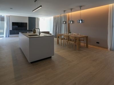 Moderne keukeneiland met geïntegreerde spoelbak en kookplaat, grenzend aan een eetruimte met houten tafel en eigentijdse hanglampen. Lichtgrijze houtlook tegelvloer.