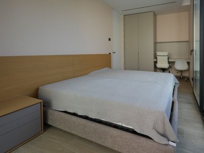 Moderne slaapkamer met lichtgrijs gestoffeerd bed, houten hoofdbord en geïntegreerd nachtkastje. Voorzien van inbouwkast, dubbele bureauhoek en grote glazen deuren.
