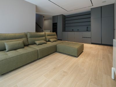 Olijfgroene modulaire bank in open woonruimte met licht houtlook tegelvloer. Donkergrijze inbouwkasten met spoelbak en planken op de achtergrond.