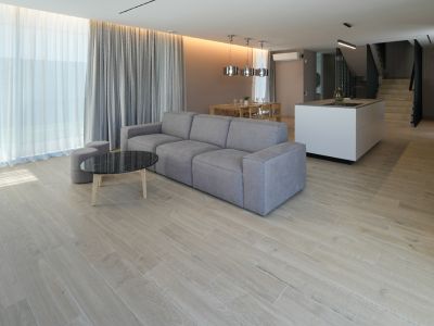 Open woonkamer met grijze modulaire bank, zwarte marmeren salontafel en houtlook tegelvloer. Grote ramen met transparante en grijze gordijnen.