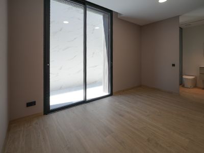 Ruime kamer met houtlook tegelvloer en lichte muren. Grote zwart omkaderde glazen schuifdeuren openen naar een buitenruimte met witte marmerlook tegels. Geïntegreerde badkamer zichtbaar.