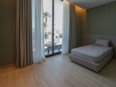 Slaapkamer met eenpersoonsbed, lichte houtlook vloer en olijfgroene muren. Grote ramen met dubbele gordijnen bieden uitzicht op de straat en palmbomen.