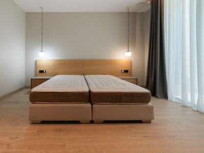 Slaapkamer met tweepersoonsbed op gestoffeerde bedframes, licht houten hoofdbord met geïntegreerde nachtkastjes en twee moderne hanglampen. Groot raam met gordijnen.