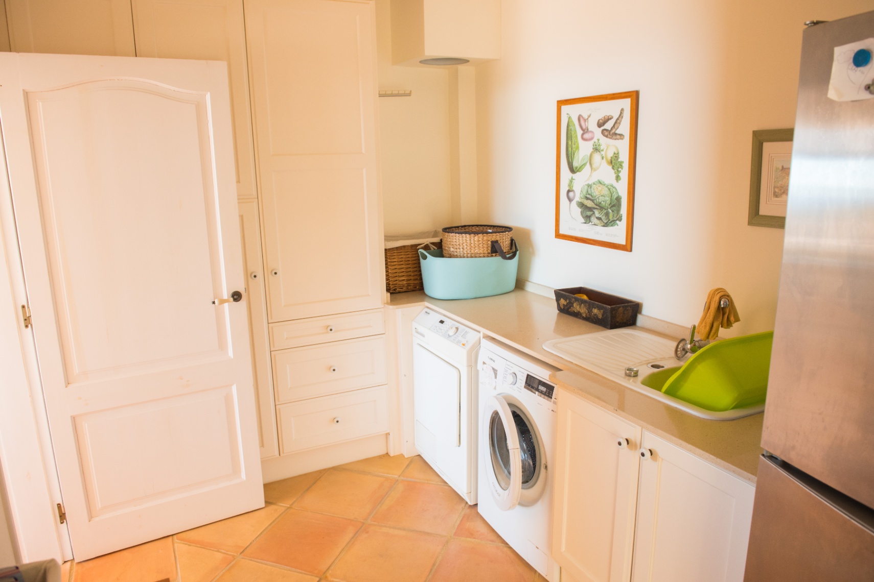 Functionele bijkeuken voorzien van witte inbouwkasten, wasmachine, droger en een klassieke terracotta tegelvloer.